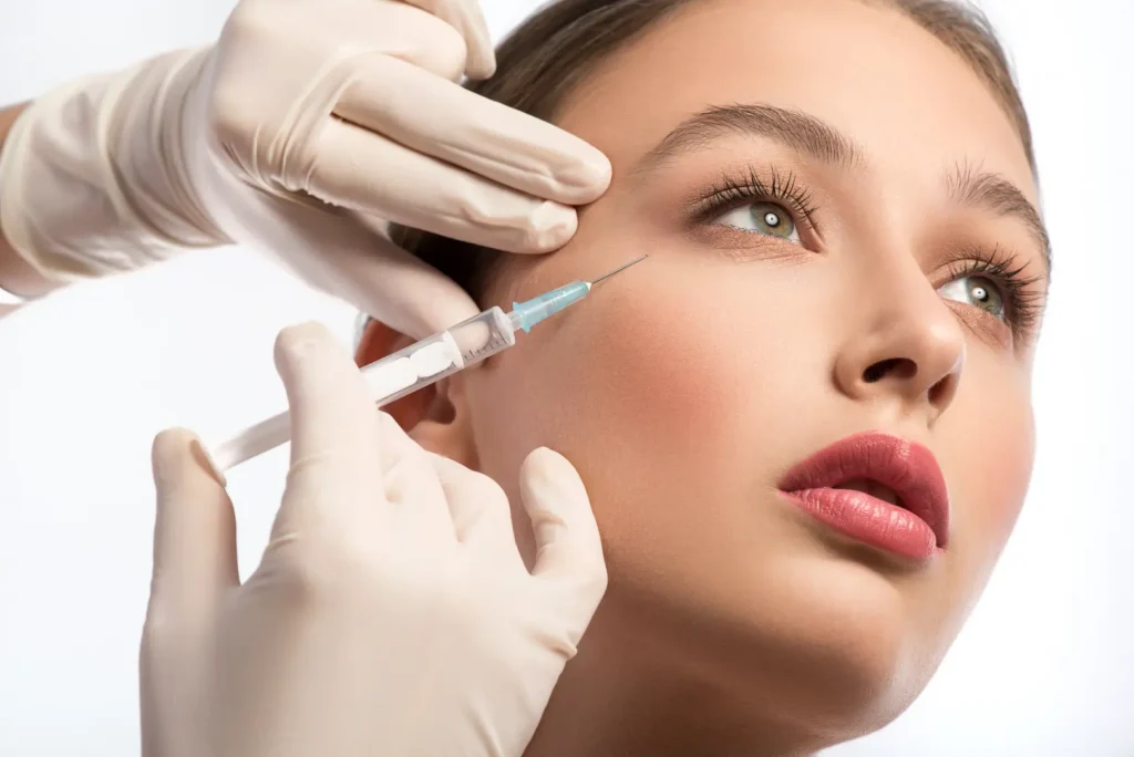 Gloved hands position a syringe near a person's cheek while gently supporting the skin during a facial injection.