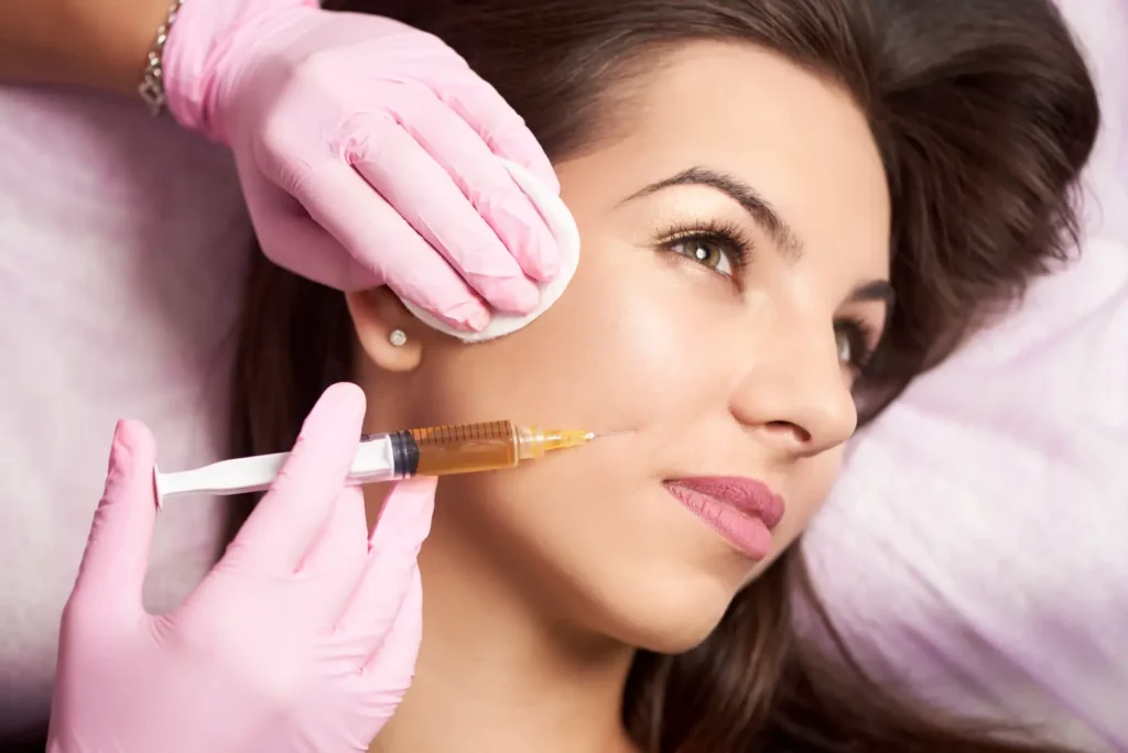 Gloved hands hold a syringe near a person's cheek while a cotton pad rests against the side of the face.