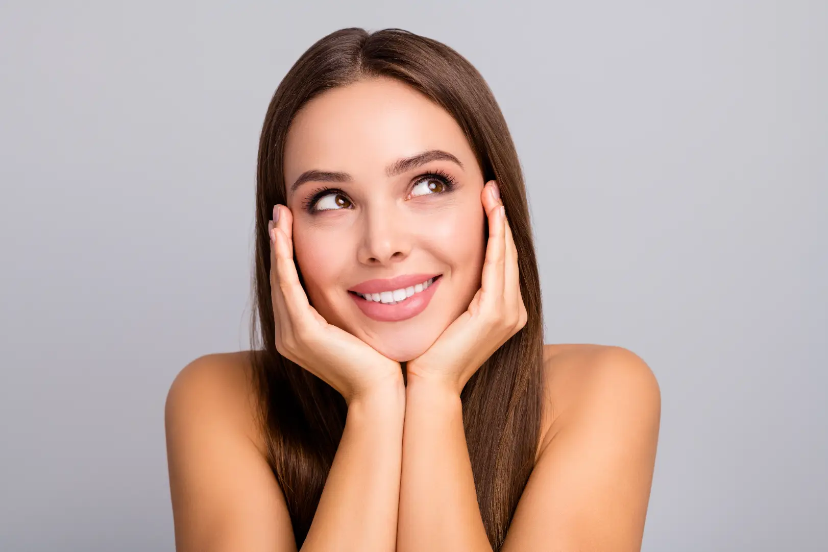 Smiling woman rests her face in both hands while looking upward against a plain gray background.