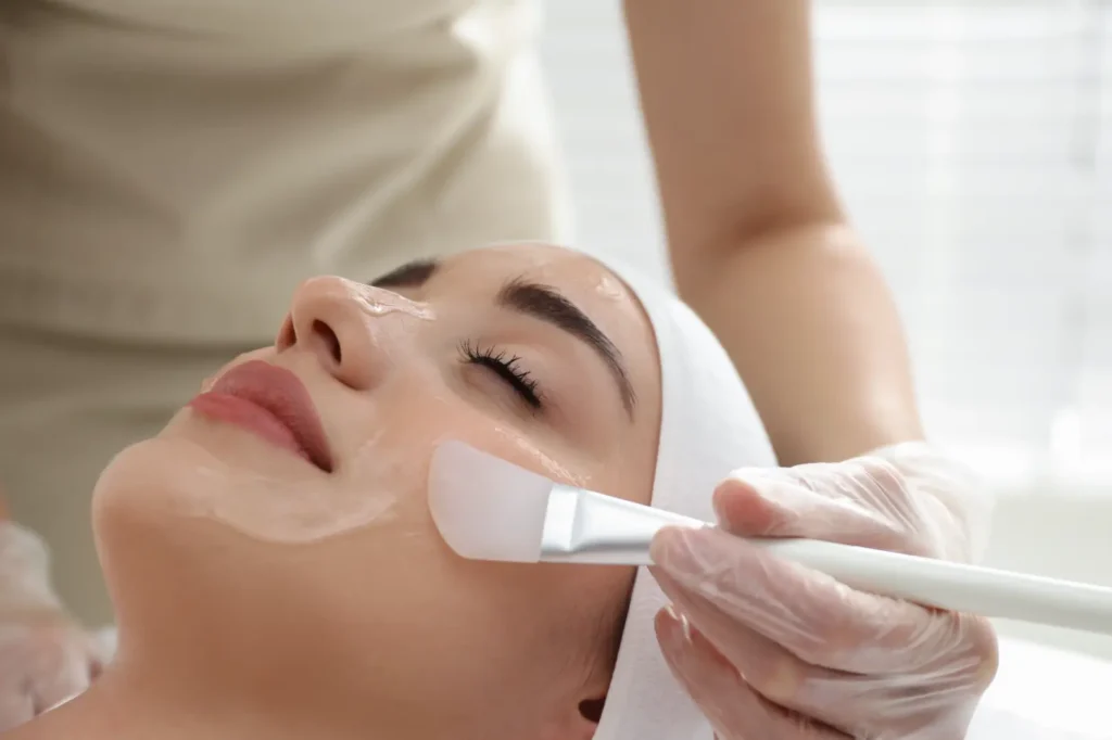 Gloved hand applies a facial mask with a flat brush to a person's cheek while they lie on a treatment bed.