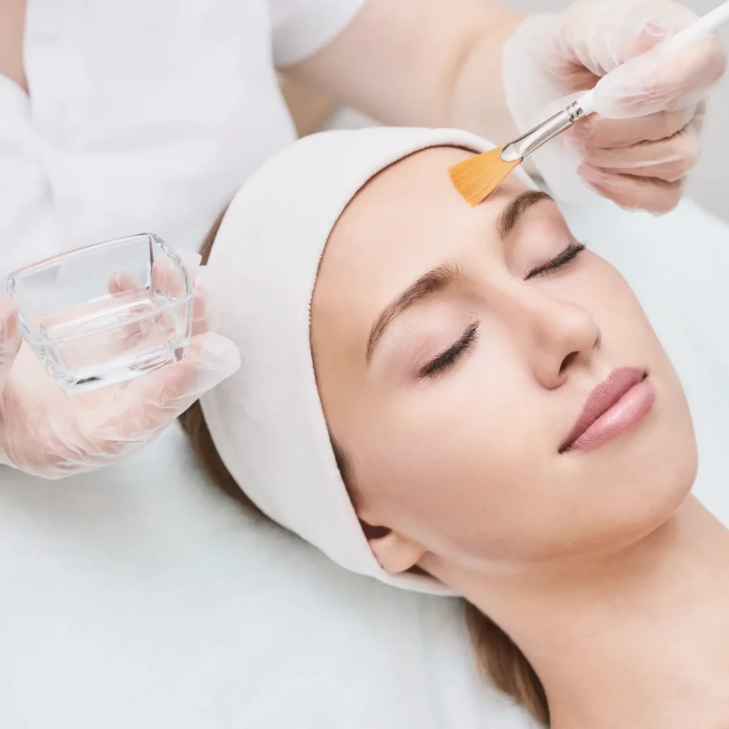 Practitioner uses a fan brush to apply a facial solution to a person's forehead while holding a glass bowl.