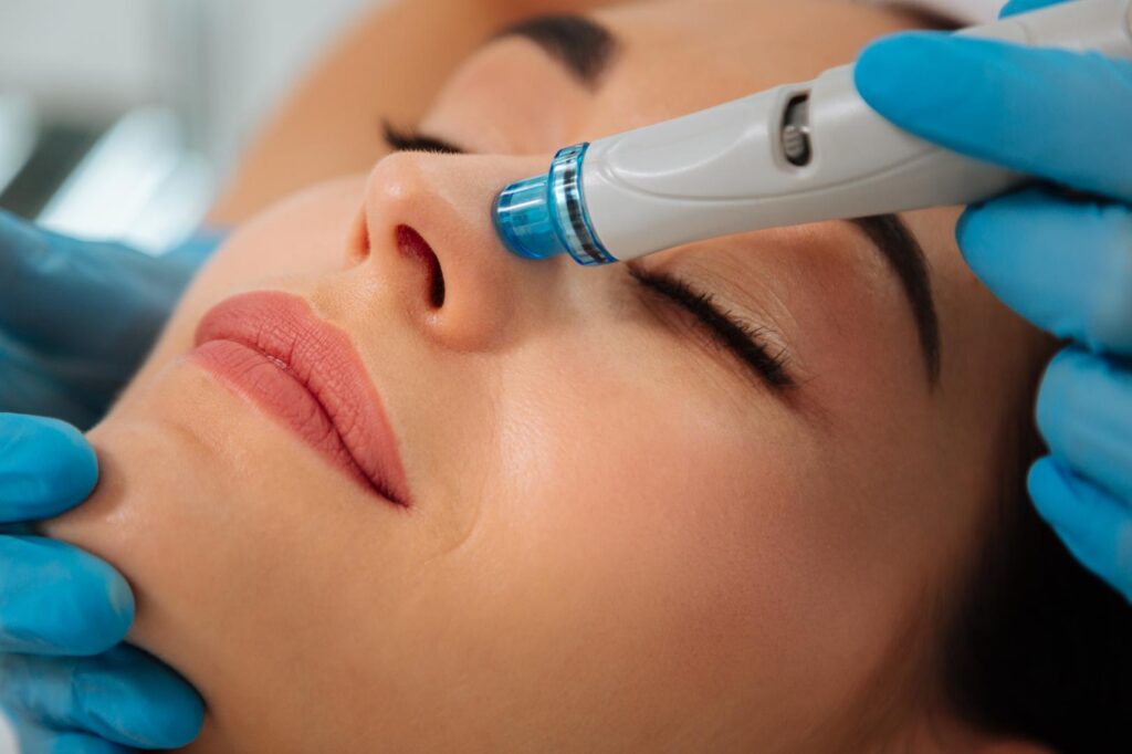 Gloved hands position a HydraFacial device on the side of a person's nose during a facial treatment.