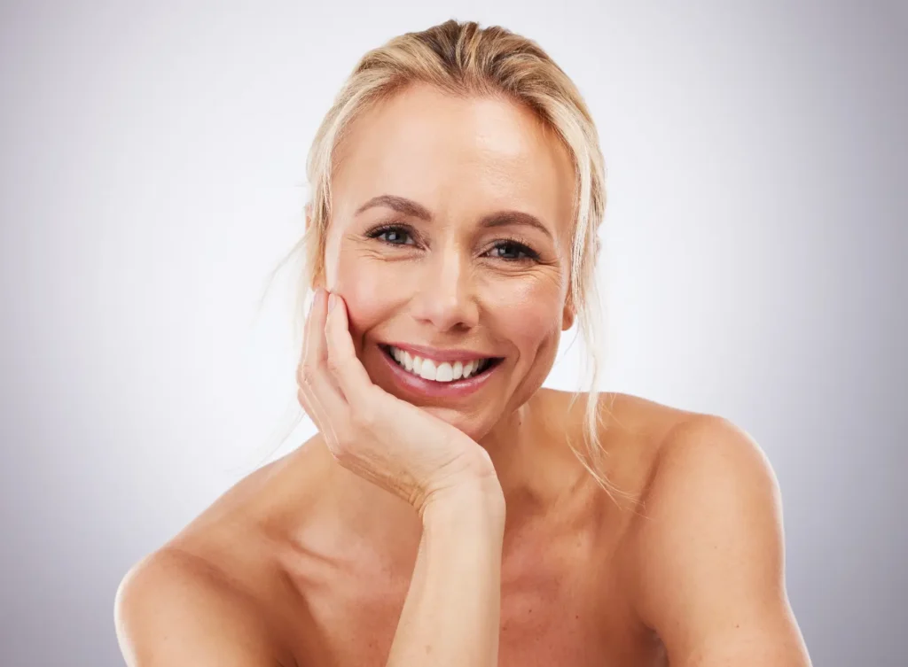 Smiling woman rests her face on one hand while looking toward the camera against a plain background.