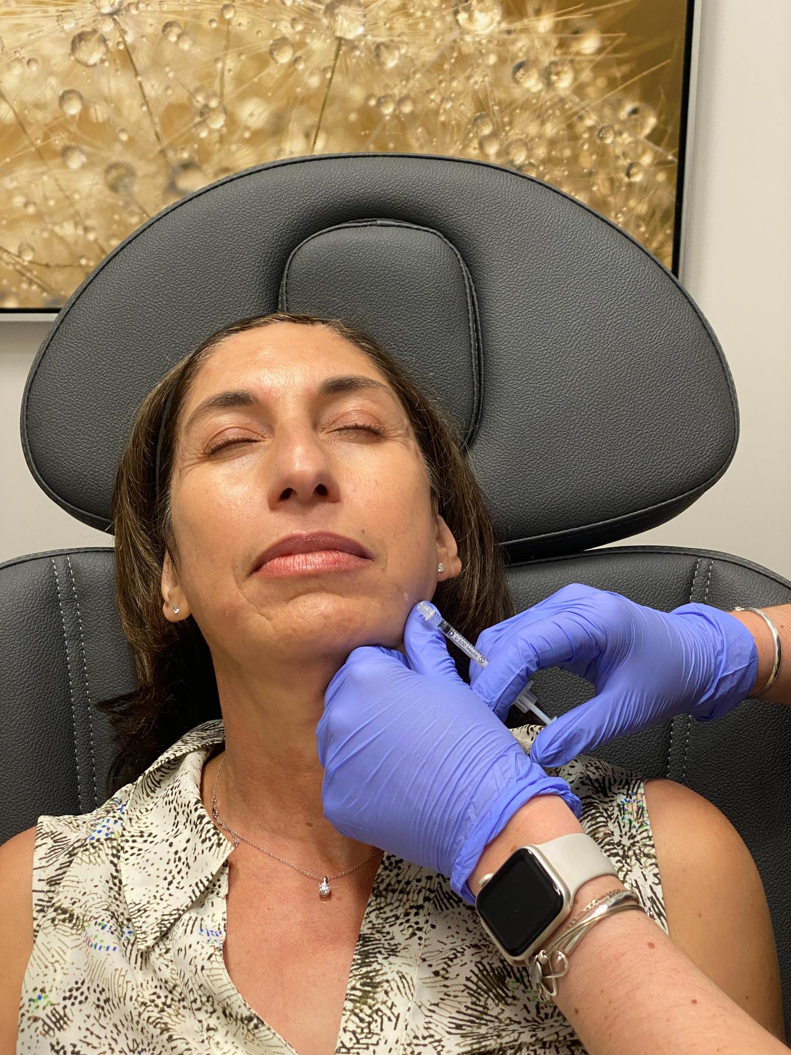 Gloved practitioner holds a syringe near a reclining woman's chin while supporting the side of her face.
