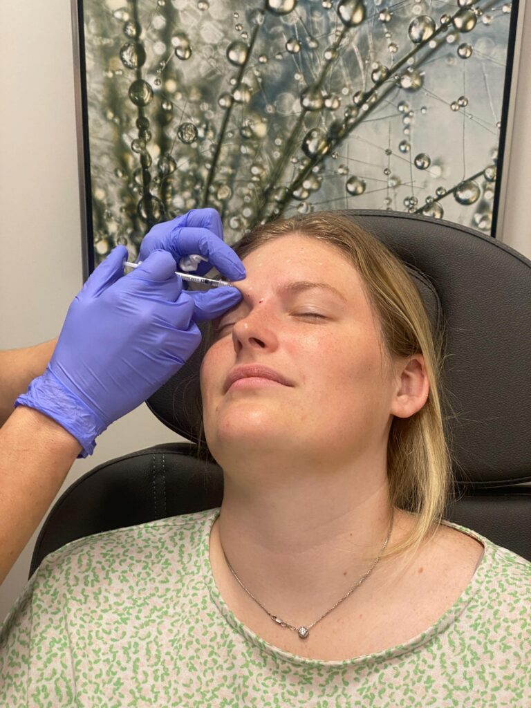 Gloved practitioner holds a syringe near a reclining woman's forehead as she rests with her eyes closed.