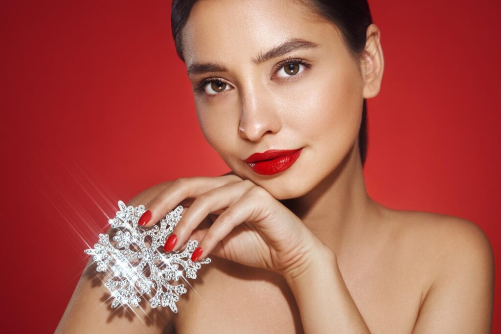 Woman with red lipstick and matching nails holds a jeweled snowflake ornament against a red background.
